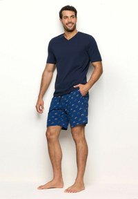Tricou navy cu decolteu în V și mâneci scurte, asociat cu șorturi albastre având un mic model tematic de camping multicolor. Poziție desculță.