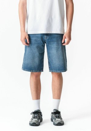 Férfi kék farmer rövidnadrágban, fehér bokazokniban, szürke és fekete sportcipőben, valamint fehér pólóban, fehér háttér előtt állva.