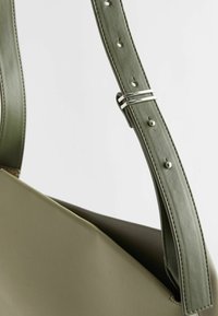 Olivengrüne Handtasche aus Leder mit glatter Textur, schräger Form und breitem Tragegurt, der mit silbernen Akzenten und Löchern zur Anpassung versehen ist.