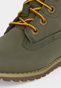 Botte en cuir vert olive avec des lacets rayés jaune et rouge passant à travers des œillets métalliques et une semelle en caoutchouc robuste.