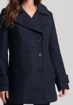 Femme portant un caban bleu marine à double boutonnage avec de larges revers et des poignets boutonnés, visible des épaules jusqu'à mi-cuisse.