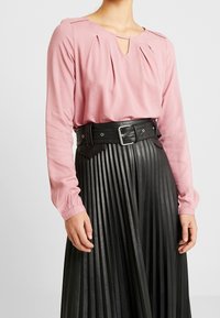 Blusa rosa a maniche lunghe con dettagli plissettati e un piccolo scollo a chiave, abbinata a una gonna nera plissettata in finta pelle e una cintura larga.