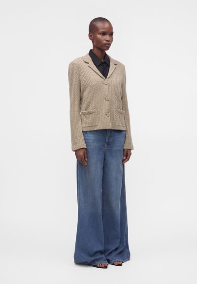 Personne portant un cardigan beige à torsades boutonné sur une chemise noire et un jean bleu à jambes larges avec des sandales noires à bout ouvert sur fond blanc.