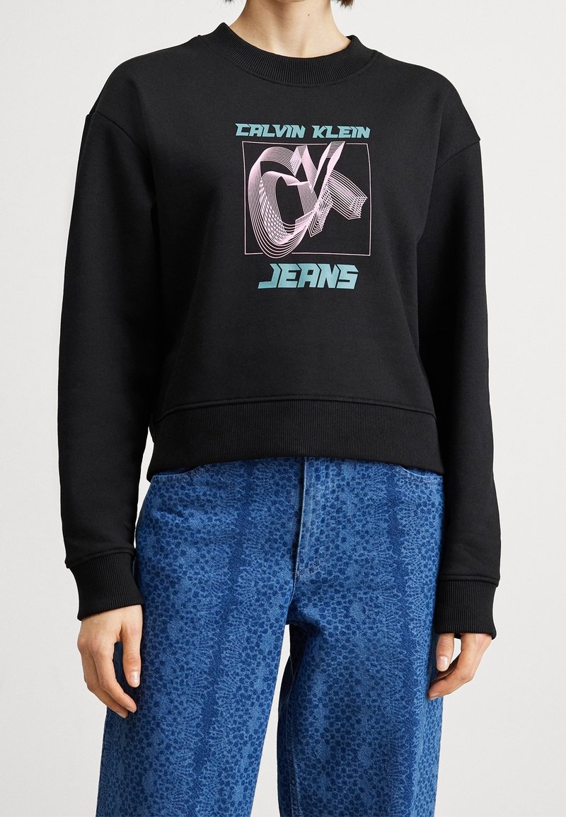 Personne portant un sweat-shirt Calvin Klein Jeans noir avec un logo géométrique bleu et un pantalon bleu ample à motifs.