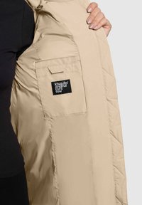 Beige színű, szigetelt dzseki gyémánttömörített textúrával, belső zsebbel és fekete címkével, amelyet kinyitva tartanak.