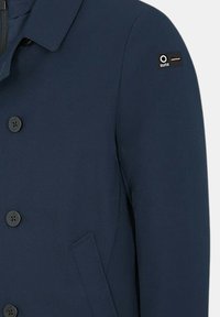 Cappotto blu navy con chiusura a bottoni, colletto, tasca laterale inclinata e piccolo patch nero del brand sulla parte superiore della manica.