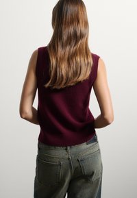 Top de punto sin mangas color burdeos con cuello acanalado, combinado con unos vaqueros de tirantes altos de color azul que cuentan con bolsillos traseros y una etiqueta de cuero.