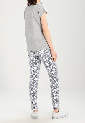 Osoba ubrana w szary krótki rękaw, białe spodnie w czarny diamentowy wzór i białe trampki, stojąca na prostym jasnym tle.