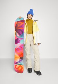 Snowboard met een kleurrijk ontwerp en "SNOW" in grote letters. Het model draagt een gele en witte jas, een mosterdkleurige coltrui, beige broek en een blauwe beanie.