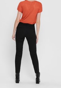 Kobieta w dopasowanej pomarańczowej bluzce z krótkim rękawem, czarnych wąskich spodniach i czarnych botkach za kostkę, stoi tyłem do widza.