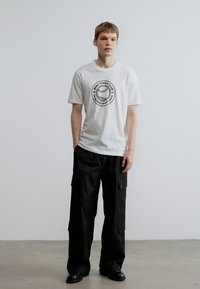 Balti medvilniniai marškinėliai su juodu apskritu teniso logotipu, derinami su juodomis krovimosi kelnėmis su daug kišenių. Lygus audinys ir laisvas siluetas.