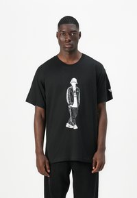 Μαύρη μπλούζα με κοντό μανίκι που διαθέτει λευκό γραφικό απεικόνισης ενός ατόμου με καπέλο και αθλητικά ρούχα. Διαθέτει στρογγυλή λαιμόκοψη και είναι φτιαγμένη από βαμβακερό υλικό.