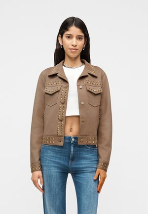 Donna con giacca corta marrone con borchie sopra un top corto bianco e jeans blu, in piedi davanti a uno sfondo chiaro e uniforme.