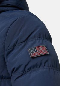 Mørkeblå polstret jakke med tekstureret stof, karakteristisk skuldersøm og et syet mærke af det amerikanske flag på overarmen.