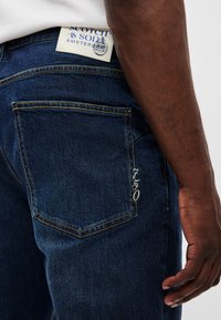 Blugi din denim albastru închis, cu o croială clasică, cusături galbene contrastante și un mic logo brodat pe buzunarul din spate.
