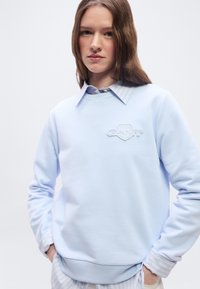 Νεαρή γυναίκα με μακριά καστανά μαλλιά, φοράει ανοιχτό μπλε φούτερ πάνω από ριγέ πουκάμισο με γιακά, τα χέρια στις τσέπες, ουδέτερο φόντο.