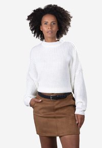 Frau mit lockigem Haar, die einen weißen Strickpullover und einen braunen Wildlederrock mit schwarzem geflochtenem Gürtel trägt, posiert mit einer Hand in der Tasche.