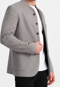 Blazer grigio a spina di pesce con colletto alla mandarino, quattro bottoni e due tasche frontali. Accenti neri sui bottoni e texture del tessuto liscia.