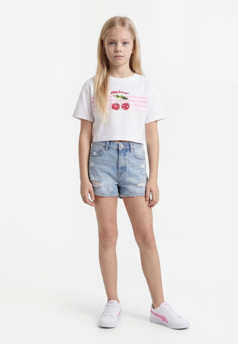 Niña joven de pie con camiseta blanca corta con cerezas, shorts de mezclilla azul claro y zapatillas blancas con detalles en rosa.