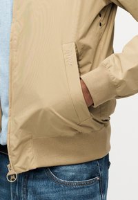 Μπεζ μπουφάν Barbour με ριμπ μανσέτες και τελείωμα, χέρι στην πλαγιαστή πλαϊνή τσέπη, φορεμένο πάνω από ανοιχτό μπλε πουλόβερ και μπλε τζιν.