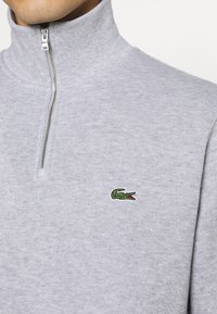 Grauer Sweatshirt mit Reißverschluss, hohem Kragen, gestrickter Textur und kleinem grünem Krokodil-Logo auf der linken Seite.