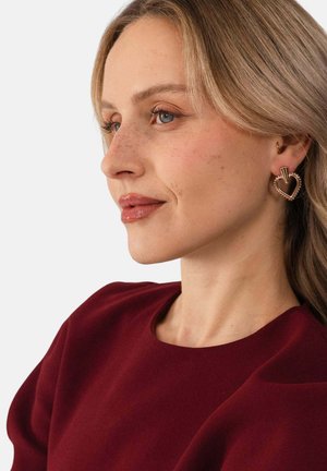 Blonde Frau mit hellen Sommersprossen, die ein burgunderfarbenes Oberteil und goldene herzförmige Ohrringe trägt und leicht nach links blickt.