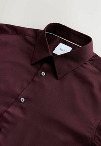 Burgundy skjorte lavet af struktureret stof med spread krave, to sorte knapper og et mærke med teksten "NEXT" indeni kraven.