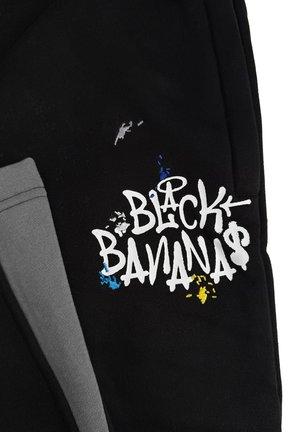 Zwarte sweatshirtstof met de tekst "BLACK BANANAS" in het wit, verfraaid met blauwe en gele verfspatten. Grijze manchet details zichtbaar.
