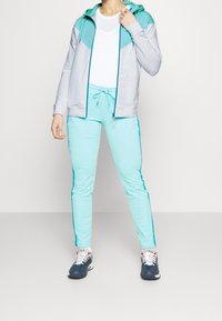 Sudadera atlética de color gris claro y aqua con cierre frontal, combinada con joggers verde menta que tienen tiras laterales, usados con zapatillas oscuras.