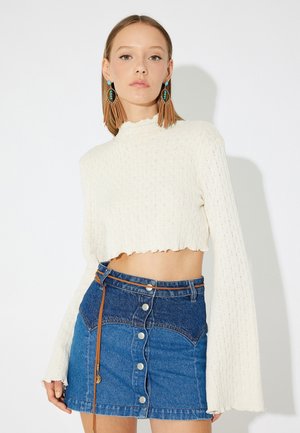 Maglione bianco cropped con un motivo testurizzato e maniche svasate, abbinato a una mini gonna in denim blu con una chiusura a bottoni e una cintura marrone.