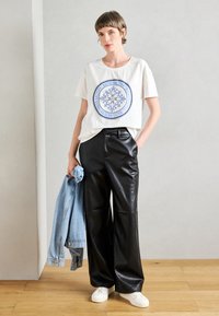 Λευκή βαμβακερή T-shirt με μπλε γραφικό σχέδιο, μαύρο δερμάτινο παντελόνι σε φαρδιά γραμμή και λευκά αθλητικά παπούτσια. Τζάκετ τζιν κρεμασμένο στον βραχίονα.