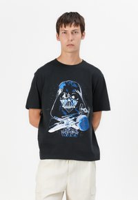 Svart bomullströja med en blå grafik av en hjälmmaskerad karaktär, ett rymdskepp och planeter, med texten "Star Wars" nedanför.