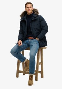Marineblauer gepolsterter Mantel mit fellgefütterter Kapuze, kombiniert mit einem marineblauen Pullover, blauen Jeans und hellbraunen Schnürstiefeln, sitzt auf einem Holzschemel.