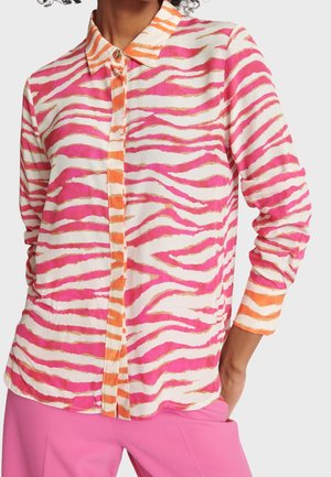 Mujer que lleva una camisa de manga larga con botones, con patrones a rayas en rosa, naranja y blanco, y pantalones rosas, con la mano apoyada en el bolsillo.