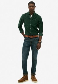 Camisa verde de botones, jeans azules, cinturón de cuero marrón y botas de cordones color beige. El modelo está de pie con los brazos relajados a los lados, en una pose casual.