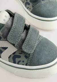 De grijs-beige kindersneakers hebben dubbele klittenbandsluitingen, gestructureerd suède en stof met een subtiele dierenprint, en witte rubberen zolen.