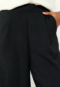 Black high-waisted trousers featuring pleats, soft texture, and side pockets. Fabric appears lightweight and slightly textured.