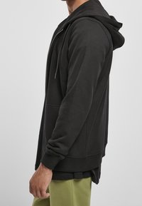 Sudadera negra con cremallera y capucha forrada, con bolsillos laterales y puños acanalados. Presenta un corte relajado y una textura de tela suave.