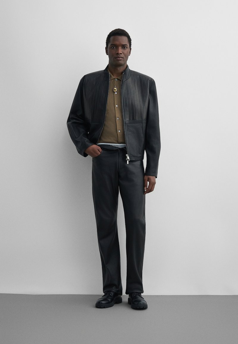 Homme portant une veste en cuir noire avec des plis verticaux, une chemise vert olive, un pantalon en cuir noir et des chaussures noires, debout contre un mur blanc.