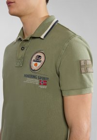 Oliwkowa polo z kołnierzykiem, z dekoracyjną naszywką i haftowanym napisem. Zawiera paski na kołnierzyku i rękawie.