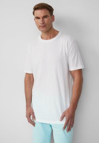 Witte katoenen t-shirt met een ronde halslijn en korte mouwen, voorzien van een subtiel logo nabij de borst. Draagt met lichtblauwe broek.