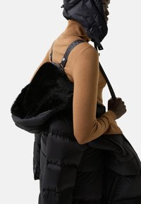 Veste en duvet noire avec un intérieur doux en fourrure noire et des sangles ajustables, portée sur un col roulé ajusté marron. Présente des détails en métal brillant.