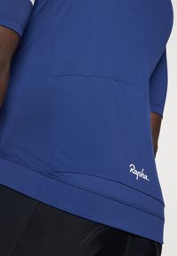 Maillot bleu à manches courtes en tissu doux avec une poche arrière et le logo blanc "Rapha." à l'ourlet. Pantalon noir partiellement visible en dessous.