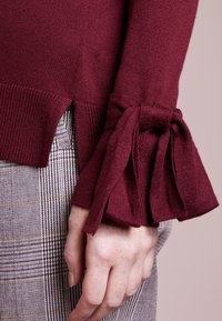 Maglione bordeaux a maglia con orlo spaccato e polsini decorati con fiocco, abbinato a pantaloni a quadri grigi con accenti gialli. Tessuto liscio, design aderente.
