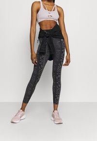 Reggiseno sportivo viola chiaro con dettagli a taglio, abbinato a leggings neri con un motivo testurizzato e logo. Scarpe da ginnastica rosa chiaro.