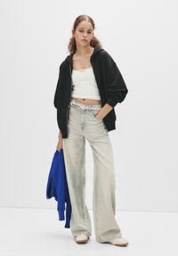 Zwarte zip-up hoodie, witte crop top, lichtgrijze wijde broek, met een zilverkleurige riem, houdt een blauwe sweater vast, crèmekleurige schoenen.