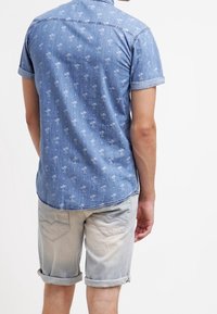 Camicia in denim a maniche corte di colore azzurro chiaro, con una leggera stampa di palme e maniche arrotolate. Abbinata a pantaloni corti in denim chiaro con orlo arrotolato.