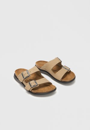 Beige toremmede sandaler med justerbare sølvspænder, brune ruskindssåler og sorte såler på hvid baggrund.