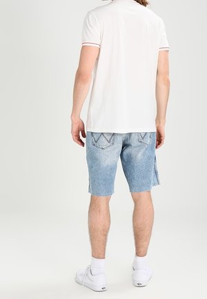 Camisa polo blanca de manga corta con acento rojo en el cuello, combinada con pantalones cortos de mezclilla azul claro con un acabado desgastado y costuras distintivas en los bolsillos traseros.