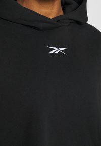 Haut noir en tissu doux, doté d'une poche kangourou à l'avant, d'une capuche avec cordon de serrage, et d'un logo blanc brodé sur la poitrine.